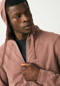 Pink hooded zip-up sweatshirt with a smooth texture, featuring a silver zipper and drawstrings. Worn over a black shirt.