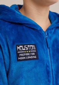 Zila flīsa kombinezons ar kapuci, ar rāvējslēdzēju priekšā un uzšuvēm ar tekstu "HOUSTON PREPARE FOR MOON LANDING" ar teksturētu apdari.