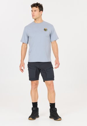 Mand stående iført lysegrå t-shirt med lille logo, mørke cargoshorts, sorte sokker og sorte vandresko på en ensfarvet hvid baggrund.