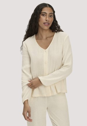 Frau mit langen lockigen Haaren, die ein cremefarbenes, strukturiertes Oberteil mit Knöpfen und passende lockere Hosen trägt, posiert mit verschränktem Arm.