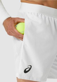 Colocando a mano una pelota de tenis amarilla en el bolsillo lateral de unos pantalones cortos atléticos blancos con un logo de ASICS.