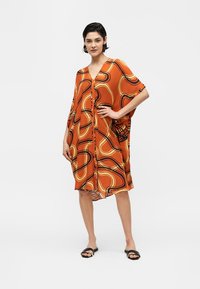 Γυναίκα στέκεται με το χέρι στη μέση, φοράει χαλαρό, πορτοκαλί φόρεμα μέχρι το γόνατο με αφηρημένα μαύρα και κίτρινα κυρτά σχέδια και μαύρα σανδάλια slide.
