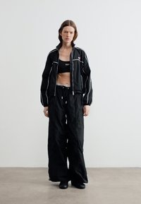 Γυναίκα όρθια, φορώντας μαύρο αθλητικό μπουφάν με λευκές ρίγες, μαύρο κοντό τοπ, φαρδιά μαύρη παντελόνα και μαύρα παπούτσια, μπροστά σε λευκό απλό φόντο.