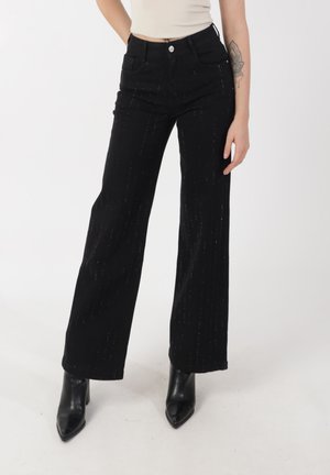 Jeans negros de pierna ancha con costuras verticales plateadas, diseño de cintura alta, cuatro bolsillos y cierre con botón, combinados con tacones negros.