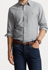 Jasnoszara koszula z kołnierzykiem, z podwiniętymi rękawami, małym logo na piersi, w komplecie z ciemnoniebieskimi dżinsami i brązowym skórzanym paskiem.