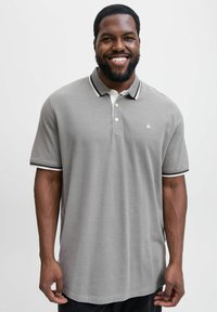 Glunderende man met een baard, gekleed in een grijze polo met zwart-witte kraag en mouwafwerking, staand tegen een effen achtergrond.