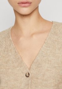 Cardigan lavorato a maglia beige con scollo a V, caratterizzato da un bottone marrone e una consistenza morbida. Sottolinea un design rilassato e accogliente.