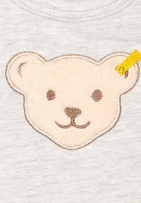 Grauer Stoff mit einem plüschigen, beigen Bärenkopf-Patch, der braune Stickereien für Augen und Nase aufweist, sowie einem gelben Akzentetikett an der Seite.