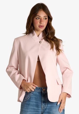 Donna con capelli castani lunghi indossa un blazer rosa chiaro abbottonato al colletto e jeans blu, posando con una mano in tasca.