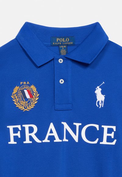 Μπλε πόλο Ralph Lauren με λευκή επιγραφή "FRANCE", χρυσό κεντημένο έμβλημα με τη γαλλική σημαία και λευκό λογότυπο παίκτη πόλο στο στήθος.