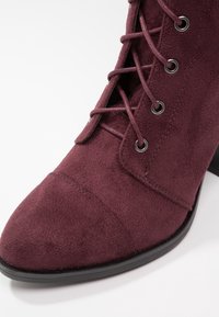 Burgundy suede ankle boot with a pointed toe, lace-up front, metal eyelets, and a black sole, featuring textured panels and stitching details.