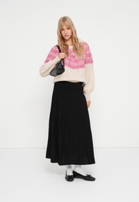 Maglione beige sovradimensionato con motivo a onde rosa, abbinato a una lunga gonna nera plissettata. Borsa nera, calzini bianchi e ballerine nere completano il look.