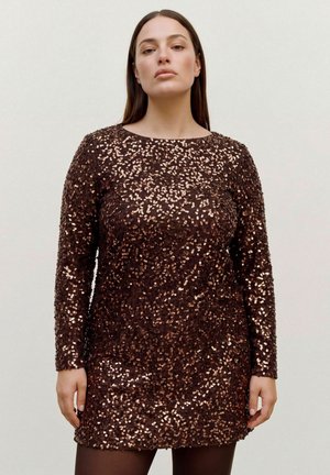 Un mannequin se tient devant un fond uni, portant une robe marron foncé à manches longues recouverte de paillettes dorées et un collant foncé.