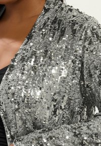 Chaqueta plateada con lentejuelas, de superficie texturizada, que presenta lentejuelas redondas superpuestas que reflejan la luz, creando un efecto brillante.