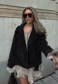 Žena s dlhými svetlými vlasmi, ktorá má na sebe tmavé slnečné okuliare, čiernu voľnú bundu, krémové šaty s volánmi a drží tmavú tašku opretú o kamennú stenou.