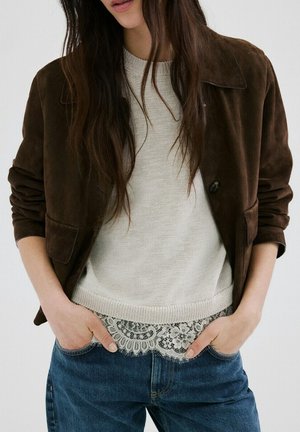 Person iført en brun ruskindjakke over en lys strikket sweater med blonder og blå jeans, hænderne afslappet i lommerne, håret løst over skuldrene.