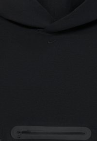 Μαύρη φούτερ Nike σε κοντινό πλάνο που δείχνει ένα μικρό κεντημένο λογότυπο swoosh και μία οριζόντια τσέπη με φερμουάρ κοντά στο γήπεδο.