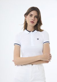 Polo shirt bianco a maniche corte con accenti blu navy sul colletto e sulle maniche. Presenta una chiusura a bottoni e un logo a forma di uccello sul petto.