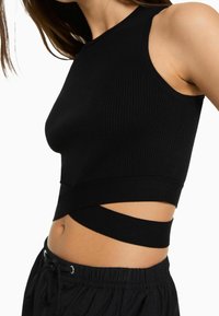 Zbliżenie na kobietę w czarnej bezrękawnikowej bluzce crop top o prążkowanej fakturze i bandzie okrążającej, zestawionej z czarnymi spodniami dresowymi z troczkami.