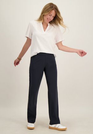 Lachende vrouw met lang blond haar die een witte blouse met korte mouwen, donkerblauwe broek en witte sneakers draagt, staat met licht opgetilde armen.