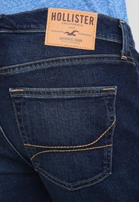 Tmavě modré džíny z denimu s hnědou koženou nášivkou s nápisem "Hollister" na pase. Obsahují dvojité švy a zadní kapsu.