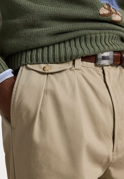 Bezs színű pliszézett chinos nadrág oldalsó zsebekkel, zöld bordás pulóverrel párosítva. A pulóver hímzett mintával és kék ingmandzsettával rendelkezik.