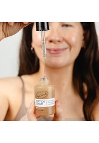 Sérum de peptides dans un flacon en verre dépoli, couleur beige, avec un bouchon compte-gouttes. L'étiquette comprend le nom de la marque et les ingrédients principaux en texte noir.