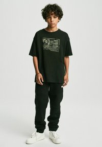 Czarny, oversize t-shirt z graficznym nadrukiem flagi i logo, połączony z czarnymi spodniami dresowymi. Białe sneakersy dopełniają całość.