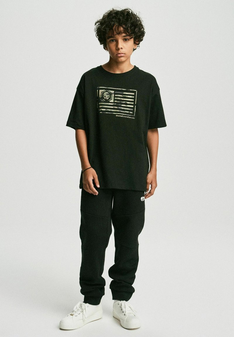 Czarny, oversize t-shirt z graficznym nadrukiem flagi i logo, połączony z czarnymi spodniami dresowymi. Białe sneakersy dopełniają całość.