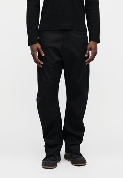 Άτομο που φοράει φαρδιά μαύρη παντελόνα, μαύρο μακρυμάνικο τοπ και μαύρα παπούτσια, στέκεται μπροστά σε απλό λευκό φόντο.