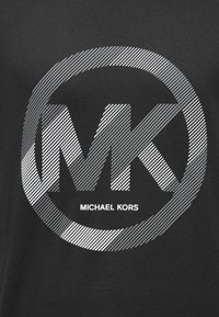T-shirt preta com um grande logótipo em riscas prateadas, apresentando as iniciais "MK" e o texto "MICHAEL KORS" em uma fonte moderna e em negrito.