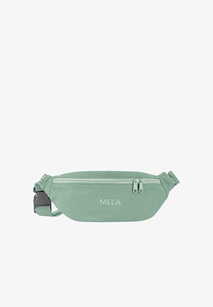 Vrouw in donkergroene trui en broek houdt lichtgroene MELA schoudertas vast tegen een witte achtergrond.