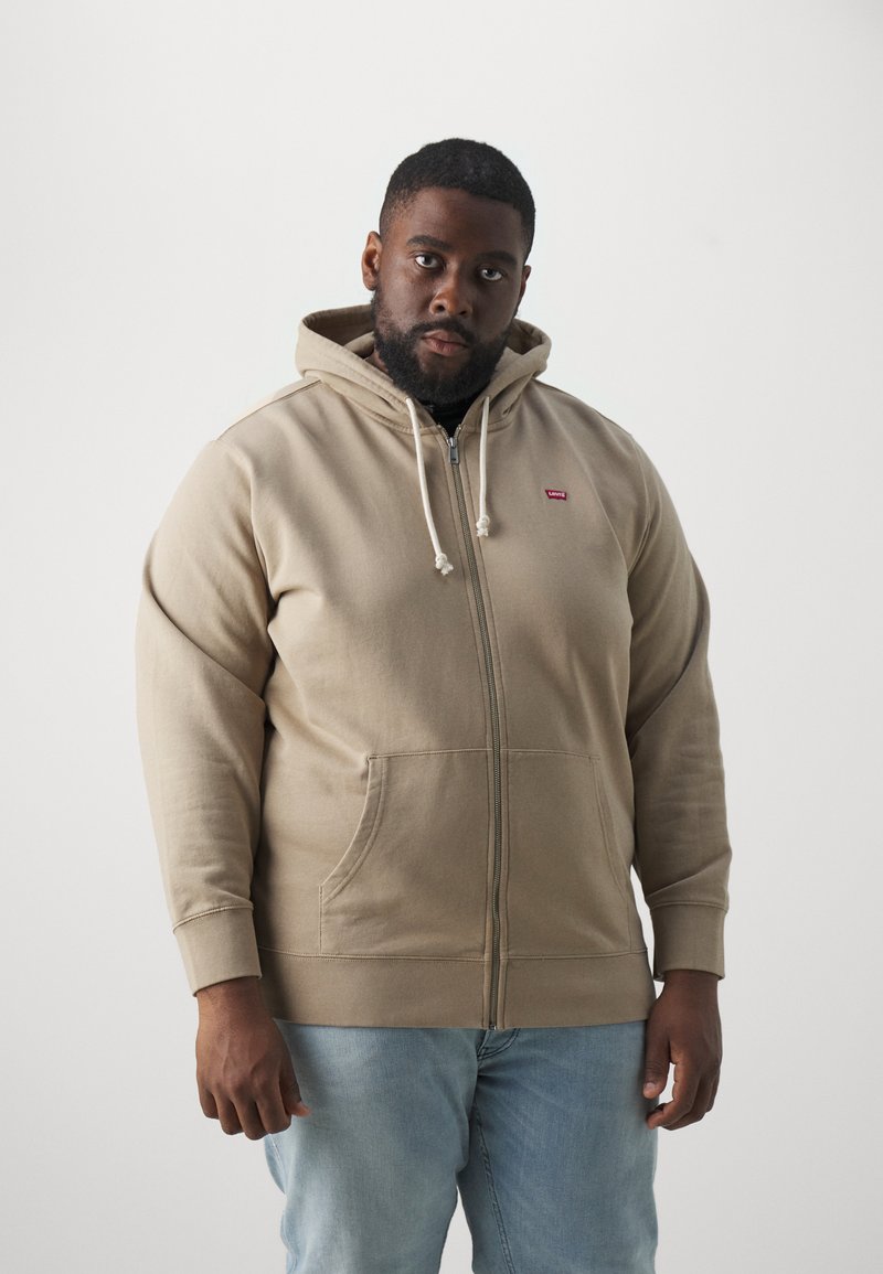 Beige Zip-Hoodie mit verstellbarem Kordelzug, vorderen Taschen, gerippten Bündchen und Logo-Patch auf der Brust. Getragen mit hellblauen Jeans.