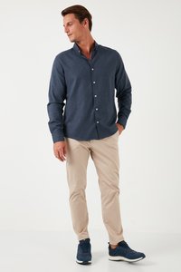 Camicia blu scuro con bottoni in un tessuto testurizzato, abbinata a pantaloni beige chiaro e scarpe da ginnastica blu scuro, esposta su uno sfondo neutro.