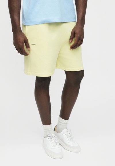 Male wearing pale yellow shorts with small "DANGAIA" logo, white crew socks, white sneakers, and a light blue shirt against a plain background.