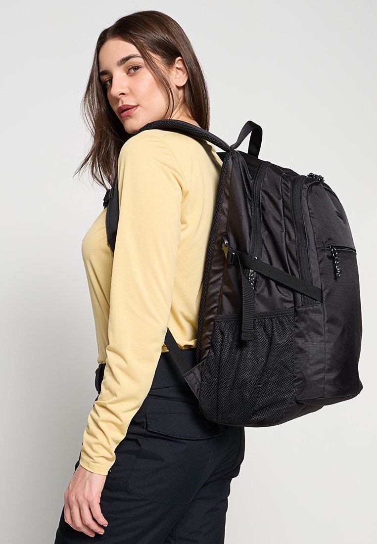 Jonge vrouw in een beige langemouwen shirt en zwarte broek, met een grote zwarte rugzak over één schouder, kijkt om.