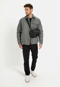 Lysegrå polstret jakke med brystlomme, sort crossover taske, sorte bukser og hvide sneakers med grå detaljer; står foran en neutral baggrund.