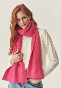 Sciarpa rosa acceso lavorata a maglia in materiale morbido, con una texture a coste e un design semplice, drappeggiata casualmente intorno al collo della modella.