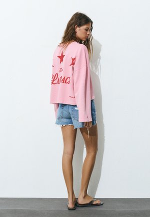 Žena stoji uz bijeli zid noseći ružičastu majicu dugih rukava s crvenim zvjezdicama i tekstom, traper šorceve i crne sandale.