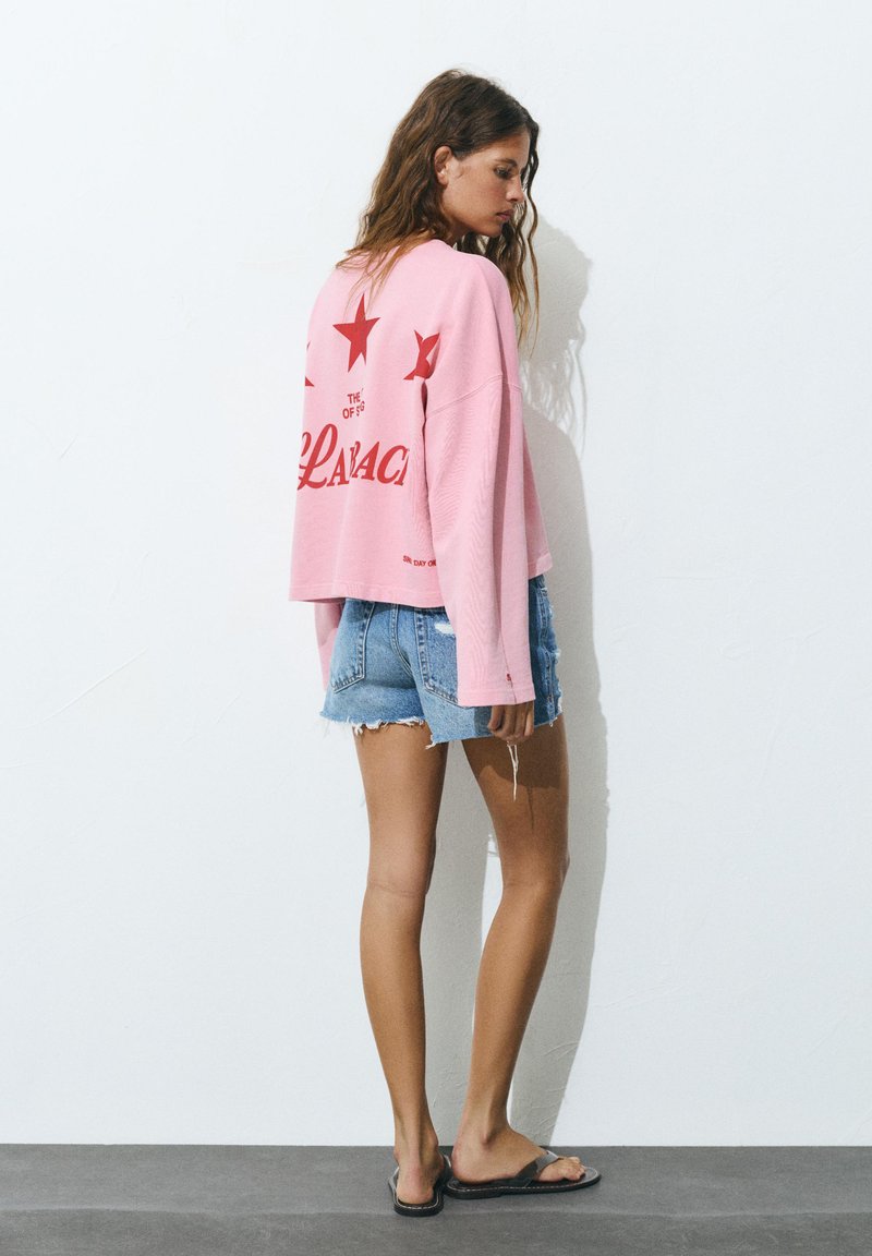 Woman standing against a white wall wearing a pink long-sleeve shirt with red stars and text, denim shorts, and black sandals.