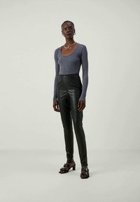 Mulher em pé, vestindo uma blusa justa cinza de manga longa, calças de couro pretas de cintura alta, sandálias pretas de salto e um colar prateado.
