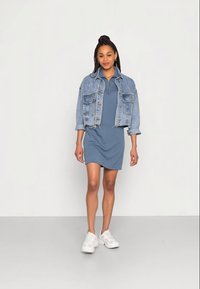 Casaco de denim sobre um vestido curto azul com colarinho com fecho. A modelo usa ténis brancos e brincos de argola de prata simples. Texturas suaves.