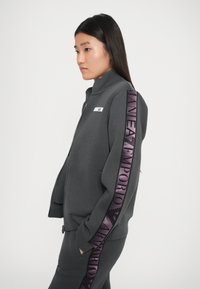 Giacca grigia con zip realizzata in materiale morbido, dotata di colletto alto, tasche laterali e una banda con logo viola lungo la manica.