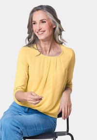 Gele longsleeve top met een ronde halslijn en geplooide details. Gecombineerd met blauwe jeans, gedragen door een zittend persoon met grijs haar.