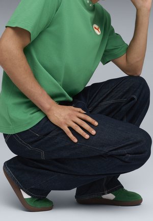 Personne accroupie portant un t-shirt vert, un jean bleu foncé et des baskets vertes avec des semelles marron sur un fond gris uni.