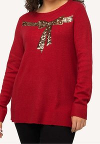 Czerwony sweter z miękkiego materiału z okrągłym dekoltem, ozdobiony kokardą z cekinów na piersi oraz ściągaczami przy mankietach i u dołu.