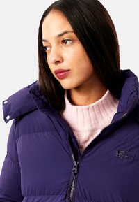 Jachetă puffer de culoare violet închis, cu închidere cu fermoar și guler înalt. Prezintă un logo Lacoste pe piept. Este purtată peste un pulover roz deschis.