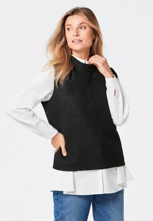 Schwarzer, gestrickter ärmelloser Pullover über einem weißen Hemd mit gewelltem Saum, kombiniert mit blauen Jeans. Merkmale einen hohen Halsausschnitt und eine gerippte Textur.