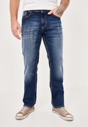 Blue denim jeans with a straight cut, light fading, contrasting stitching, five pockets, and worn with gray sneakers.