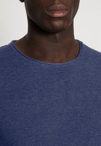 T-shirt bleu marine texturé à col rond, présentant un motif tricoté subtil et un encolure lisse. La vue en gros plan met en valeur les détails du tissu.